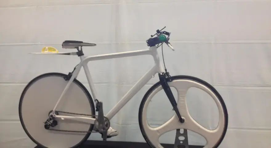 A sleek, white 3d printed bike with unique disc and tri-spoke wheels. Yellow sunglasses rest on the seat.