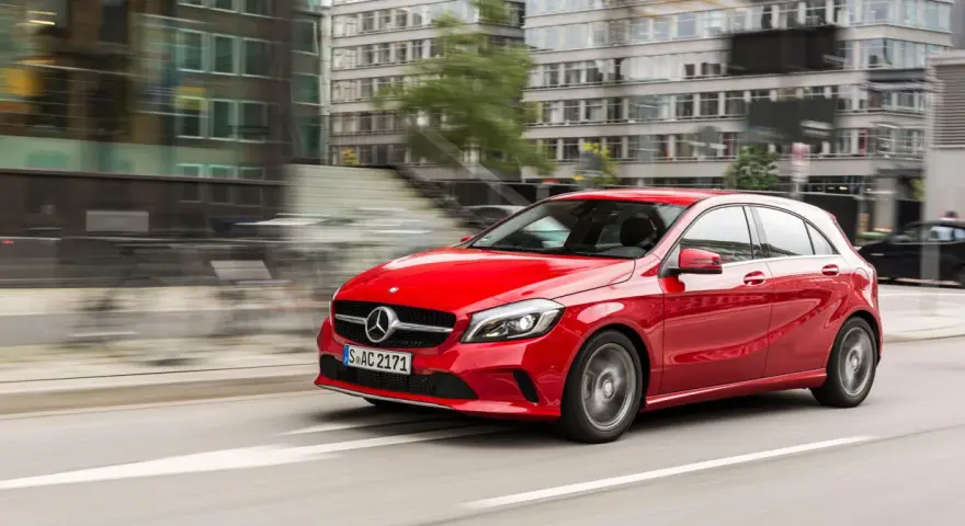 Czerwony Mercedes-Benz W176 pędzi ulicą miasta, mijając nowoczesne budynki.