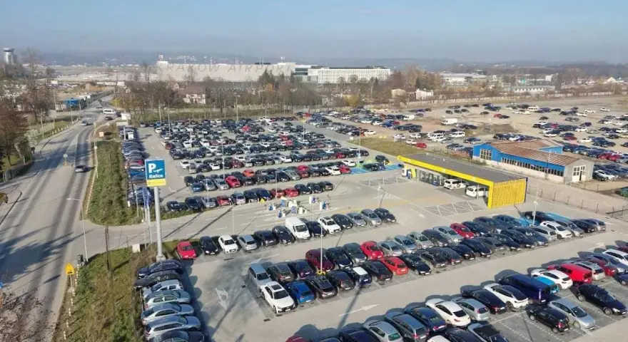 Duży parking przy krakow katowice airport. Wiele samochodów zaparkowanych w rzędach, w tle budynki lotniska.