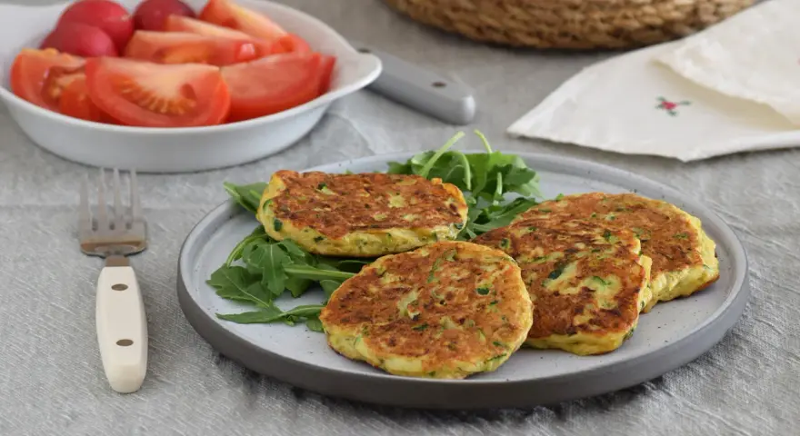 Tortitas de calabacín doradas y esponjosas, servidas sobre rúcula fresca. Acompañadas de una ensalada de tomate y rábano.