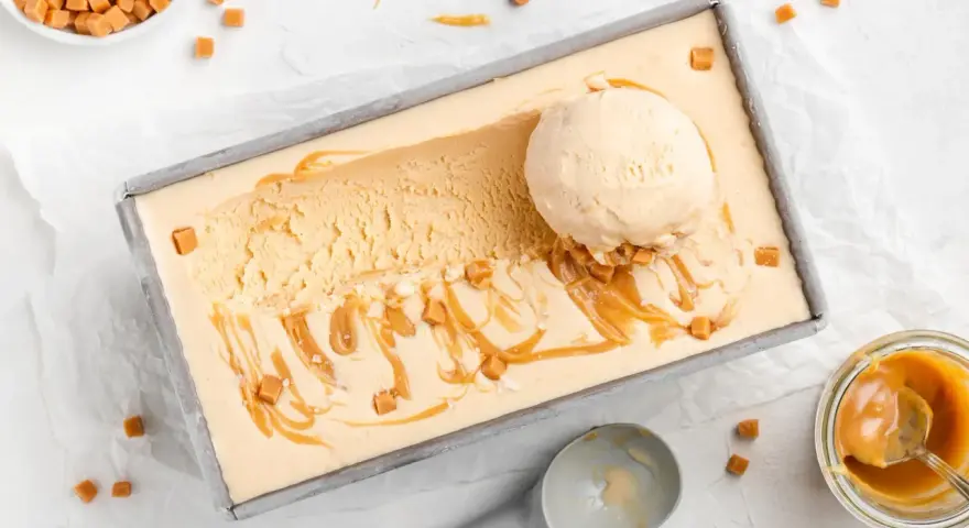 Ein Blech mit cremigem, hellbraunem Salted Caramel Eis, garniert mit Karamellsauce und kleinen Karamellwürfeln. Eine Eiskugel liegt darauf.