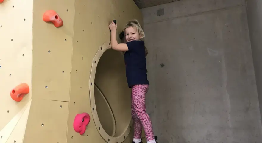 Ein fröhliches Mädchen klettert in einer Kletterhalle für Kinder. Sie lächelt und hält sich an einem Griff fest.