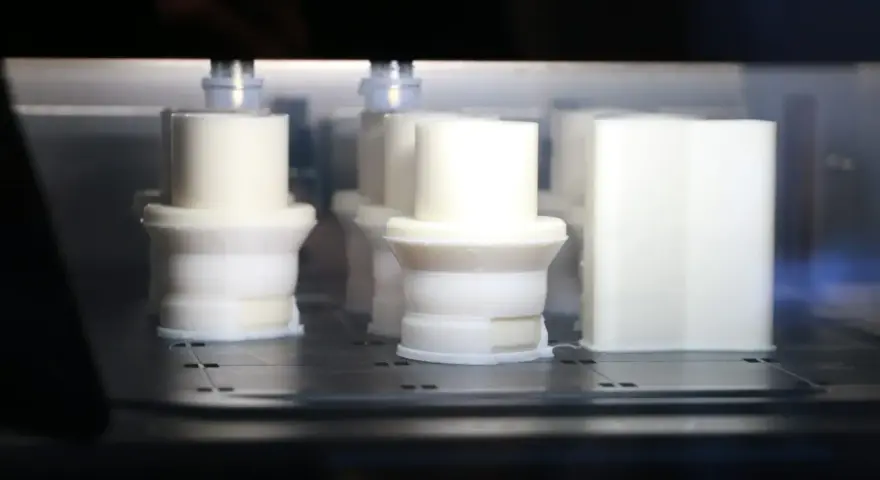 Białe obiekty w drukarce 3D, z widocznym elementem przypominającym 3d print raft, tworzą się warstwa po warstwie.