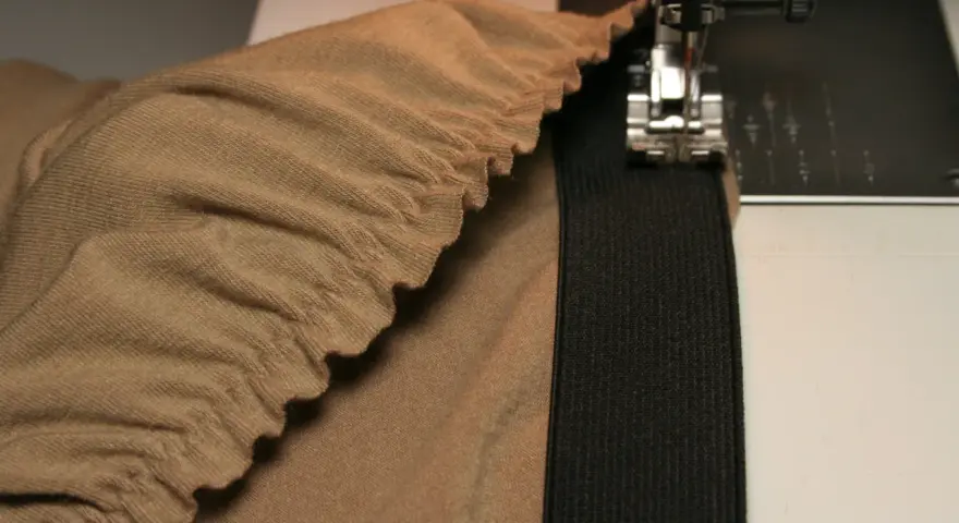 Machine à coudre en train de coudre un élastique sur un tissu beige froncé. Le pied presseur est visible, ainsi que l'aiguille.