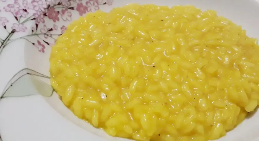 Un cremoso risotto allo zafferano, servito in un piatto bianco decorato con fiori rosa e rami verdi.