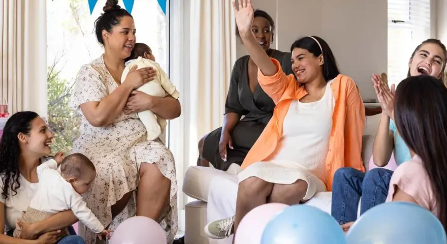 Glada kvinnor firar en babyshower med ballonger och bebisar. En kvinna vinkar glatt.
