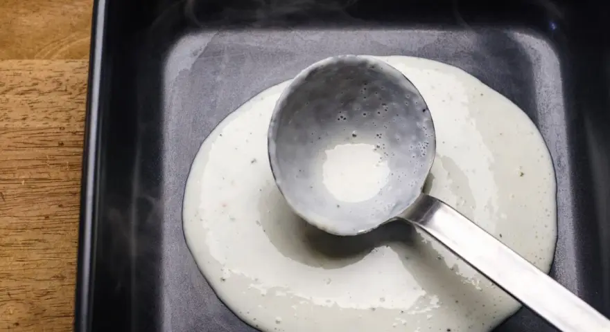 Eine Schöpfkelle verteilt cremige Bechamel-Sauce in einer schwarzen Auflaufform.