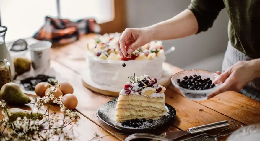 Manos expertas decoran un pastel en una mesa de reposteria y pasteleria, con frutas frescas y arándanos.