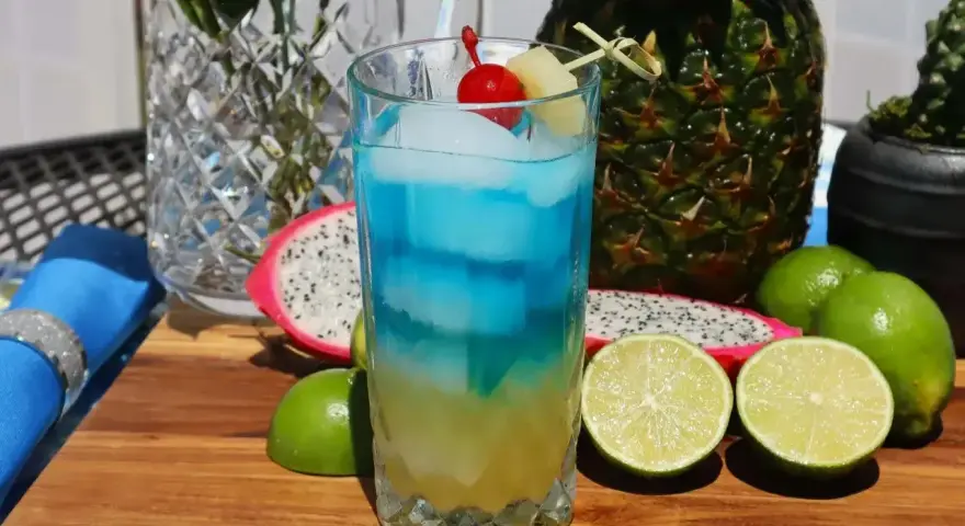 Koktajl z blue curacao alkohol, lodem, wiśnią i ananasem, otoczony owocami: smoczym owocem, limonkami i ananasem.