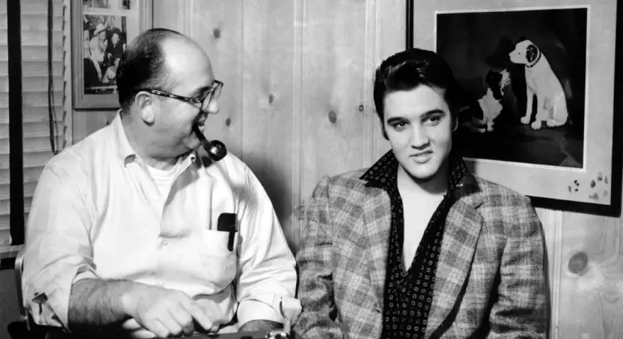 Elvis Presley und Colonel Parker, sein Manager, sitzen nebeneinander. Parker raucht Pfeife, Elvis blickt in die Kamera.