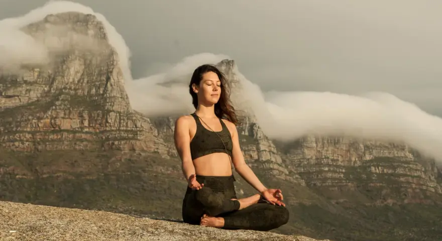 Kvinna med slutna ögon mediterar på en klippa med berg och moln i bakgrunden. Detta är en bild av vad betyder meditation.