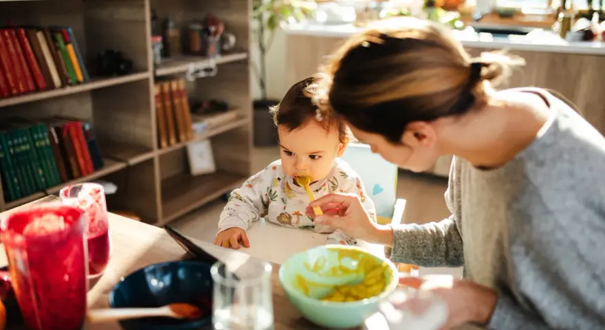 Mama karmi łyżeczką 6 miesięczne dziecko. Na stole są owoce i napoje.