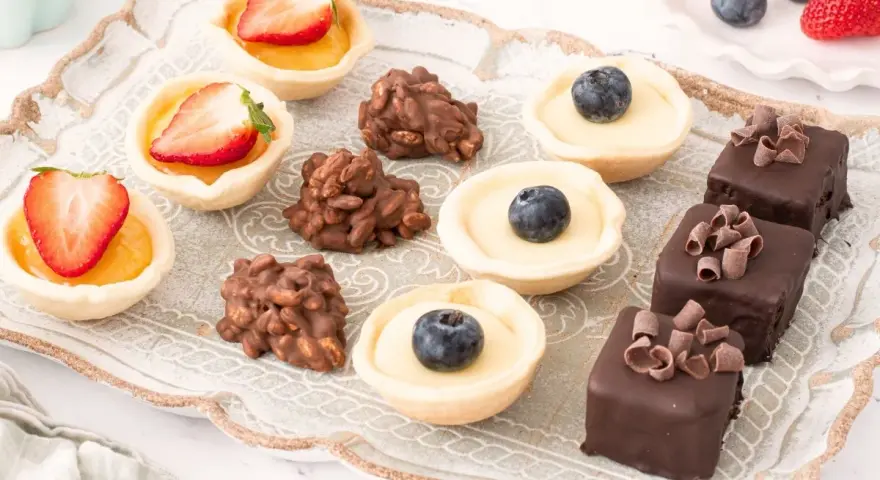Bandeja con variedad de petit fours: tartaletas de fresa y arándano, bombones de chocolate y dulces de frutos secos.