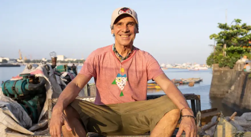 Le chanteur Mano Negra, souriant, assis sur un banc rouillé, avec un collier coloré et un t-shirt à motif poisson.