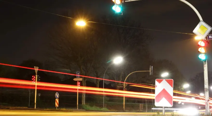 Nachts an einer Kreuzung mit roten Lichtspuren von Autos. Ampeln zeigen Grün und Rot. Ein Vorfahrtsschild und ein Richtungspfeil sind zu sehen. 132 bkat.