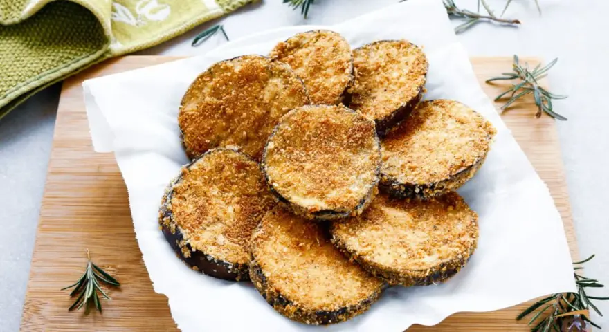 Cotolette di melanzane dorate e croccanti, servite su carta bianca con rametti di rosmarino.