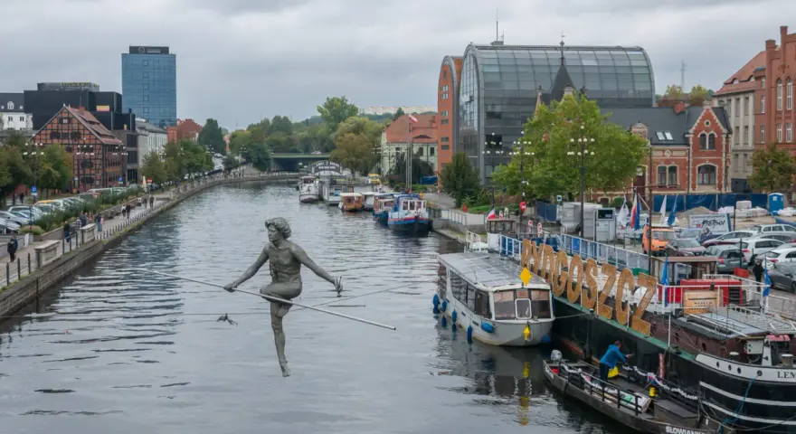 Bydgoszcz atrakcje: rzeźba na linie nad Brdą, cumujące statki i budynki nad rzeką.