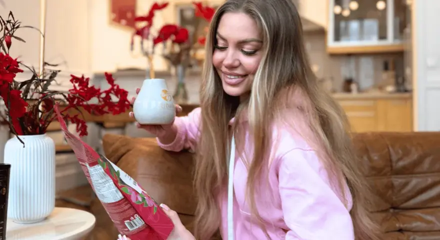 Uśmiechnięta kobieta trzyma kubek i paczkę yerba mate, przygotowując się do parzenia.