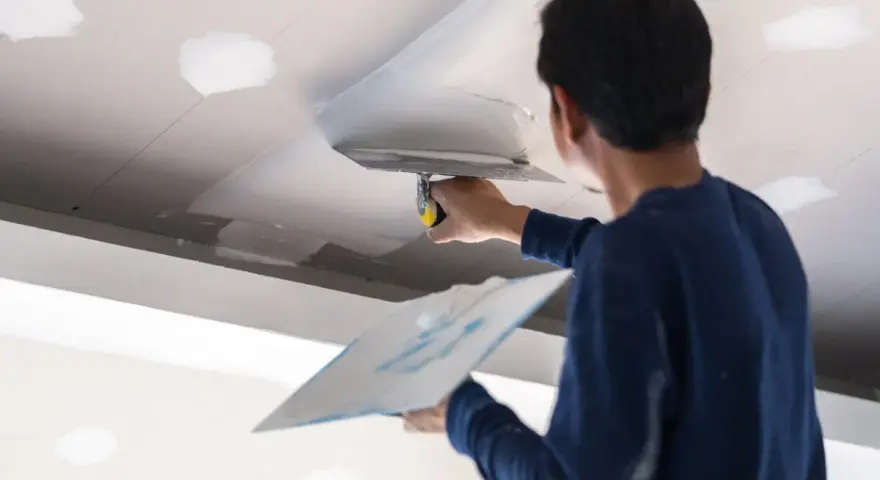 Un trabajador aplica masilla al techo, resolviendo la duda de si que se pinta primero el techo o las paredes.
