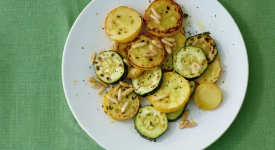 Gegrillte gelbe Zucchini-Scheiben mit Pinienkernen und Kräutern auf einem weißen Teller.