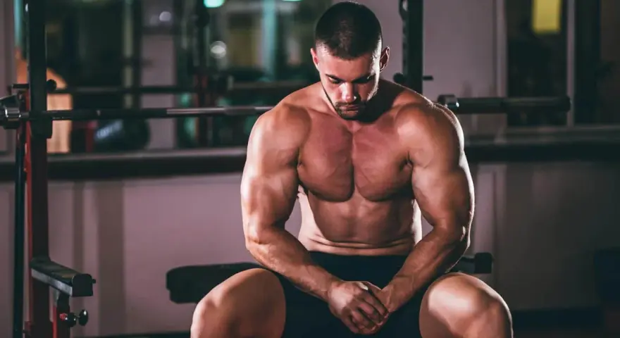 Un homme musclé, concentré, s'entraîne pour prendre du muscle rapidement. Il est assis dans une salle de sport, le regard baissé.