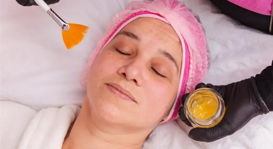 Kobieta w czepku leży z zamkniętymi oczami, a kosmetyczka przygotowuje maseczkę z miodu na twarz, używając pędzelka.