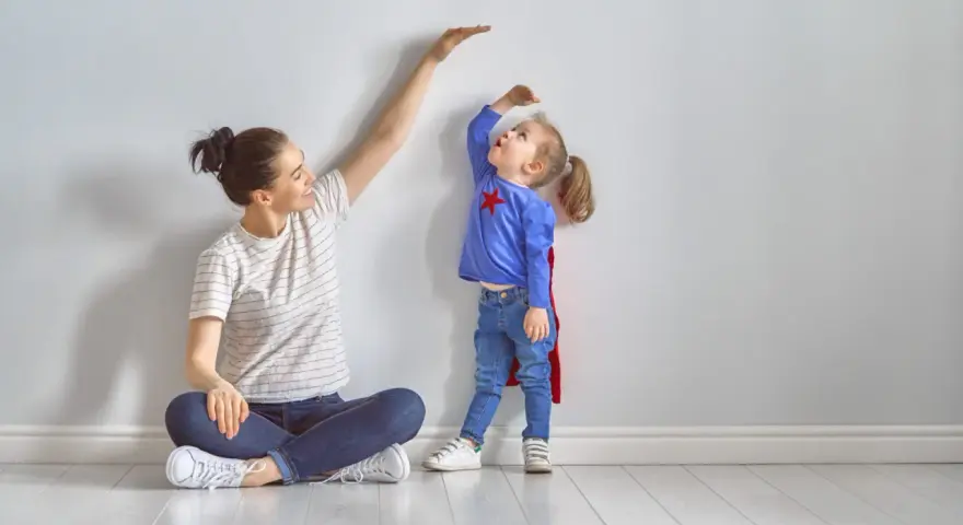 Mama mierzy wzrost córeczki w stroju superbohaterki. To właśnie wtedy dzieci rosną najszybciej, zaskakując nas każdego dnia.