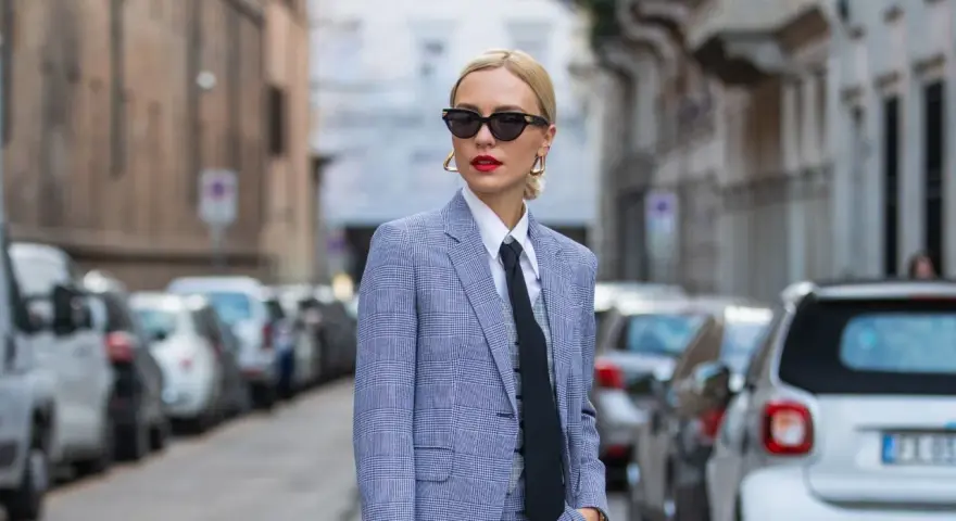 Estilo tomboy que es: mujer con traje a cuadros, camisa blanca y corbata negra, gafas de sol y labios rojos, posando en la calle.