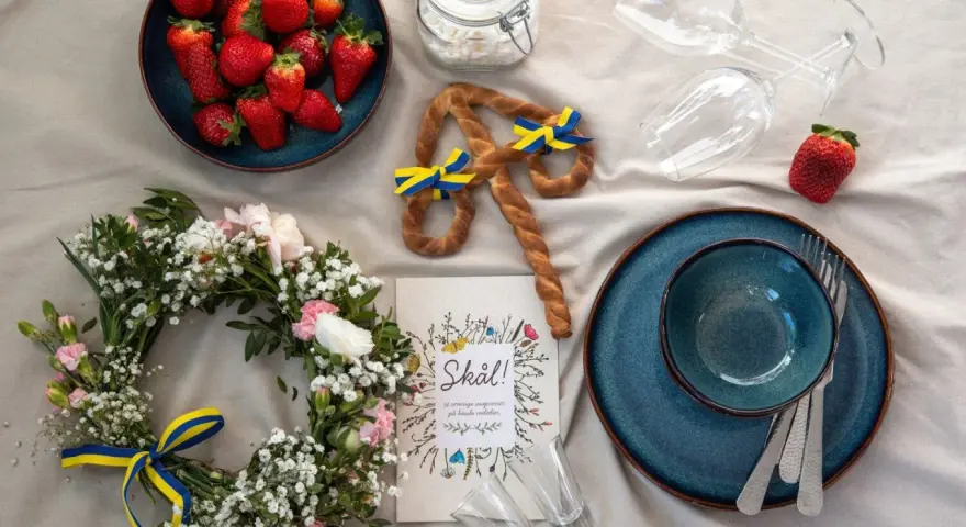 En midsommardukning med jordgubbar, blomsterkrans, en pretzel med svenska flaggor och en "Skål!"-bok. Ett personligt ljus för firandet.