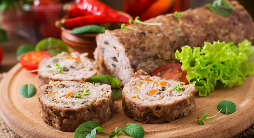 Pokrojone zrazy z mięsa mielonego z warzywami na desce, obok sałata, pomidory i papryczki chili.