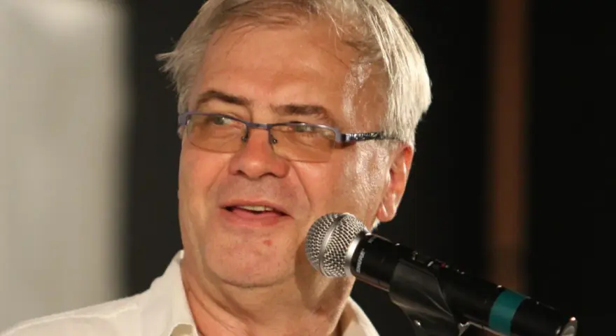 Andrzej Błaszczak, aktor, w okularach i białej koszuli, przemawia do mikrofonu.
