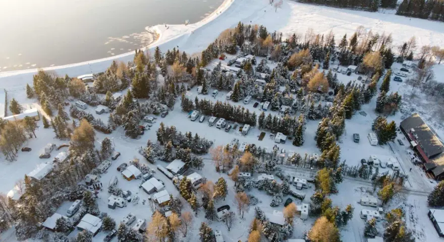Wintercamping im Erzgebirge: Wohnmobile und Wohnwagen stehen im Schnee auf einem Campingplatz am See, umgeben von verschneiten Bäumen.
