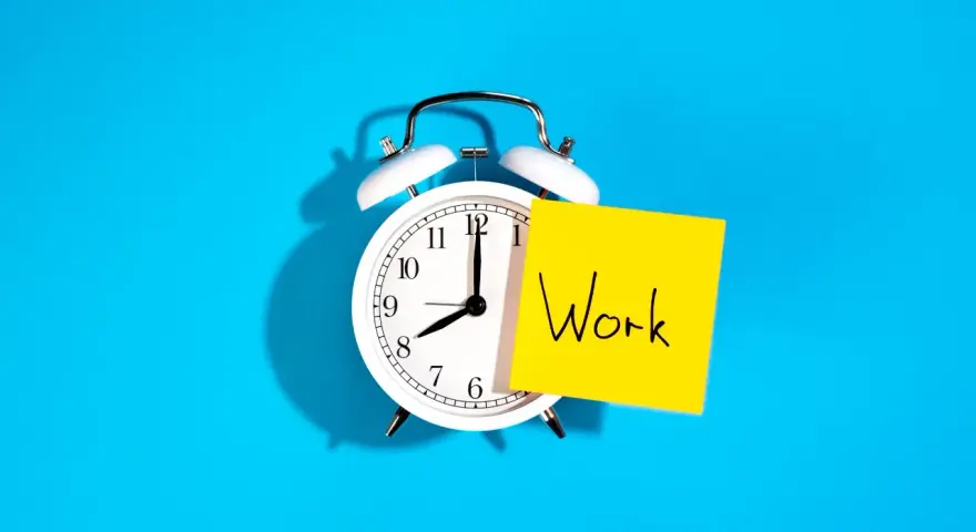Reloj despertador blanco con nota amarilla que dice "Work", recordando la importancia de la jornada laboral.