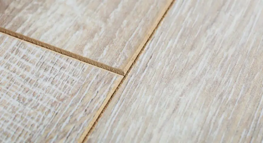 Zbliżenie na połączenie paneli podłogowych. Widoczne są dwie deski z jasnym, drewnopodobnym wzorem i wyraźnym frezem, pokazujące, jak połączyć rozchodzące się panele.