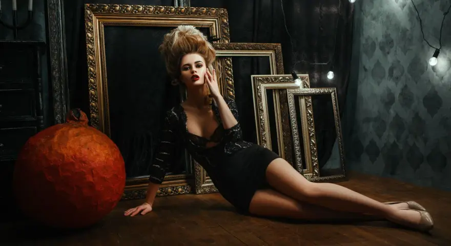 Une femme aux cheveux volumineux, dans une robe noire, pose près de cadres dorés. Un tuto Photoshop pour un look dramatique.