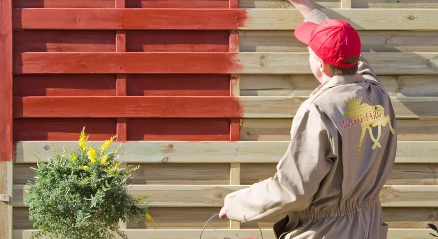 Person in beige Overall lackiert Holzzaun rot. Das geht auch ohne Schleifen, wie die Marke "Moose Färg" zeigt.
