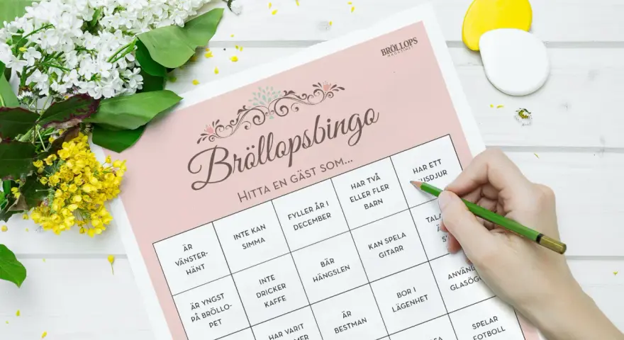 Bröllopsbingo med roliga frågor med svar. En hand med en grön penna markerar rutor på ett spelblad. Blommor ligger bredvid.