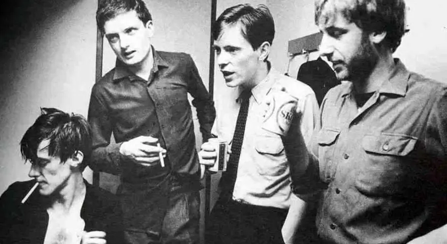 Les membres de Joy Division, Ian Curtis, Bernard Sumner, Peter Hook et Stephen Morris, posent ensemble, certains fumant, d'autres tenant des boissons.