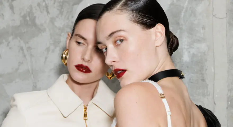 Due modelle con labbra audaci e un intenso colore rossetto, con capelli tirati indietro e orecchini dorati.