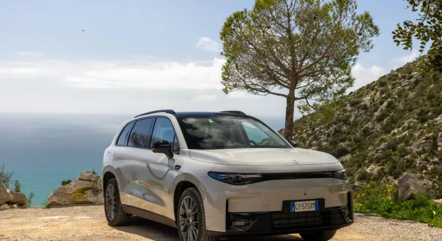 Un SUV blanco moderno, uno de los tipos de coches eléctricos, aparcado en un camino de tierra con el mar y una colina verde de fondo.