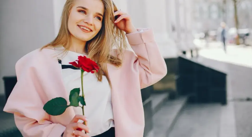 Uśmiechnięta blondynka z różą w dłoni, w różowym płaszczu. Idealna stylizacja: jak się ubrać do kina z chłopakiem, by wyglądać olśniewająco.