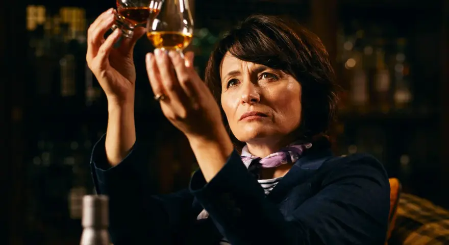 Rachel Barrie betrachtet konzentriert zwei Gläser Whisky, während sie in einer dunklen Bar sitzt.