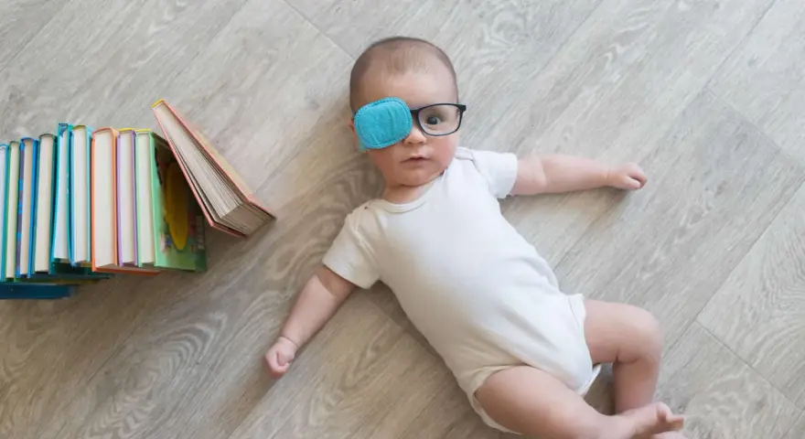 Bambino con occhiali e cerotto sull'occhio per correggere l'occhio ambliope, sdraiato vicino a una pila di libri colorati.