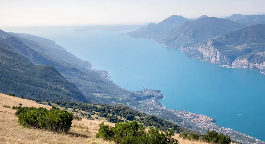 Panoramica mozzafiato del lago di Garda sponda veronese, con montagne verdi e un borgo pittoresco affacciato sull'acqua azzurra.