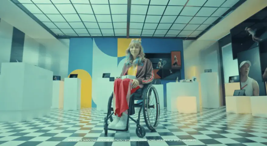 Zofia Komasa w wózku inwalidzkim, otoczona nowoczesnymi ekspozycjami, z kolorowym tłem i charakterystyczną podłogą w szachownicę.