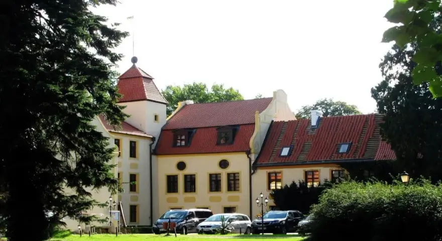 Zamek w Krokowej z czerwoną wieżą i dachami, otoczony zielenią. Przed budynkiem zaparkowane samochody.