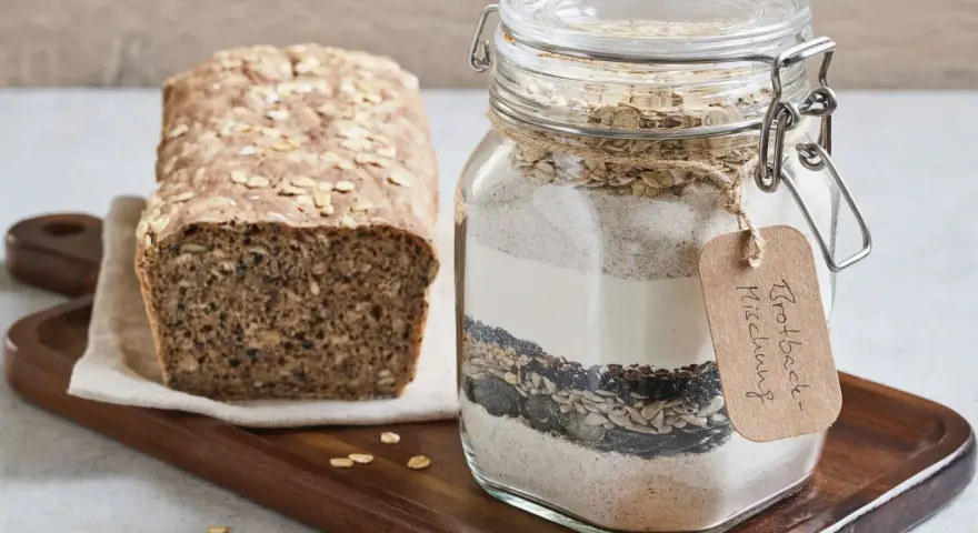 Selbstgemachte brotbackmischung im Glas, bereit zum Backen. Daneben ein frisch gebackenes Brot mit Haferflocken und Körnern.