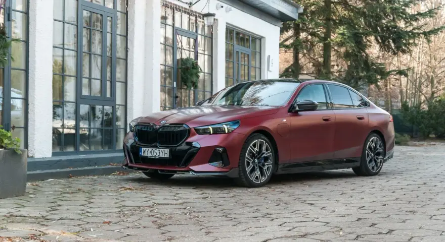 Eleganckie, matowoczerwone BMW 550e z czarnymi akcentami stoi na brukowanym podjeździe przed budynkiem z dużymi oknami.