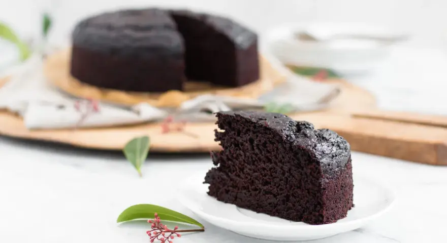 Ein saftiges Stück veganer Kuchen liegt auf einem Teller, daneben ein ganzer Schokoladenkuchen.