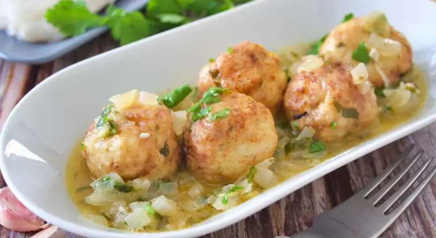 Cuatro albóndigas de merluza doradas en una salsa cremosa con cebolla y perejil, servidas en un plato blanco.